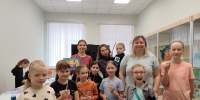 Группа детей и взрослых в зале мастерской: дети держат кисти, на столах видны рисунки, в витрине — выставленные работы. - 168.Ru