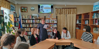 Группа школьников за столами в библиотеке; преподаватель стоит с листом на фоне книжных полок и экрана с афишей «Есенинский диктант». - 168.Ru