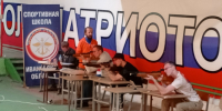 Несколько стрелков в спортивном тире сидят за столами и стреляют из пневматических винтовок на фоне большого баннера с триколором и надписью «ПАТРИОТ» и стенда «СПОРТИВНАЯ ШКОЛА Ивановской обл.» - 168.Ru