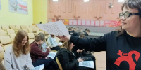 В зале школьники сидят в рядах с распечатками, на которых видны QR‑коды; ведущая протягивает лист с QR‑кодом. - 168.Ru
