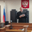 Судья в мантии за кафедрой в зале заседаний; на стене герб РФ и российский флаг; микрофоны и столы заседания на переднем плане. - 168.Ru