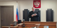 Судья в мантии за кафедрой в зале заседаний; на стене герб РФ и российский флаг; микрофоны и столы заседания на переднем плане. - 168.Ru