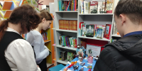 Посетители осматривают выставочный стол с тряпичными куклами в библиотеке; на заднем плане книжные стеллажи и вывеска с названием выставки. - 168.Ru