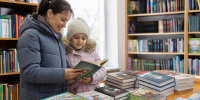 внутри библиотеки: женщина и девочка читают книгу у стола с выставленными новыми книгами; на заднем плане книжные стеллажи - 168.Ru