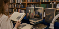 Книжная выставка: на столе раскрытые и стоящие книги с портретами и иллюстрациями, рядом посетитель, на заднем плане деревянные книжные стеллажи. - 168.Ru