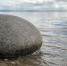 Низкий близкий ракурс: один серый береговой камень у мелкой воды, к нему подходит лёгкая ветровая рябь; камень в центре или чуть сбоку, позади размытая серо-голубая вода. - 168.Ru