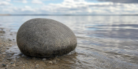 Низкий близкий ракурс: один серый береговой камень у мелкой воды, к нему подходит лёгкая ветровая рябь; камень в центре или чуть сбоку, позади размытая серо-голубая вода. - 168.Ru