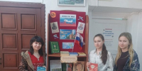 Книжная выставка на стеллаже с плакатом «Вместе мы — Россия», российским флагом и тремя молодыми посетителями у стен библиотеки - 168.Ru